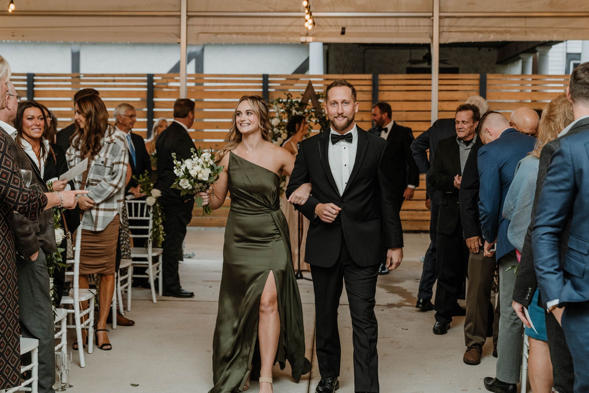 Bride and groom walk down aisle after ceremony; guests smile. Bride in green dress, groom in black tuxedo.