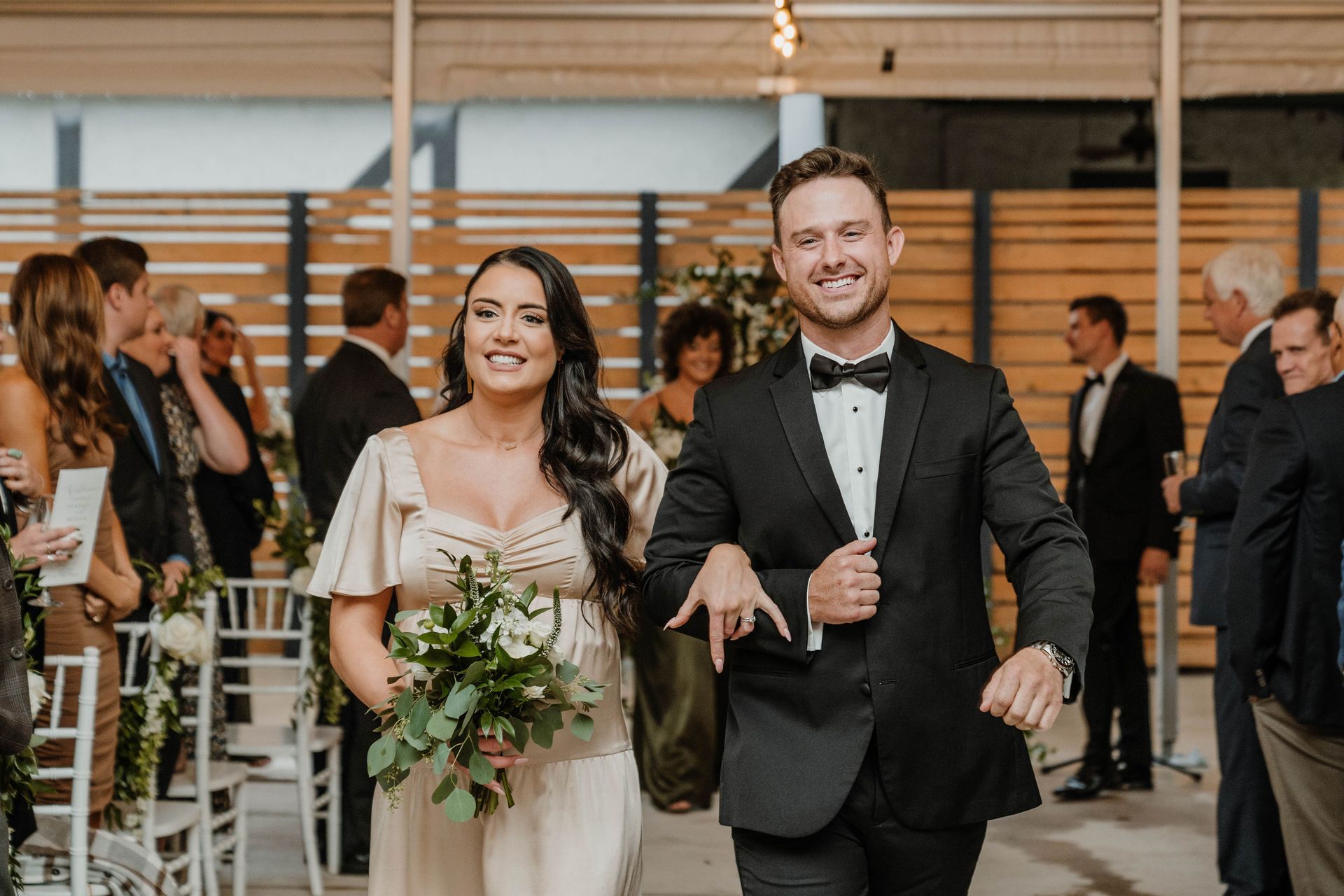 Newlyweds walking, smiling. Ceremony in modern space, man in tuxedo, woman in light dress, holding bouquet.