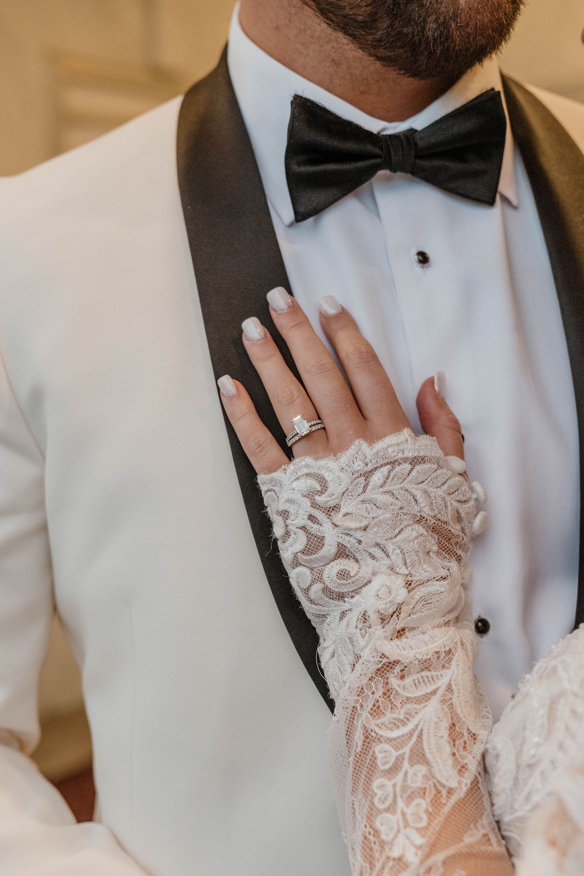 Bride's hand with ring on groom's white suit jacket with black bow tie. Lace glove detail.