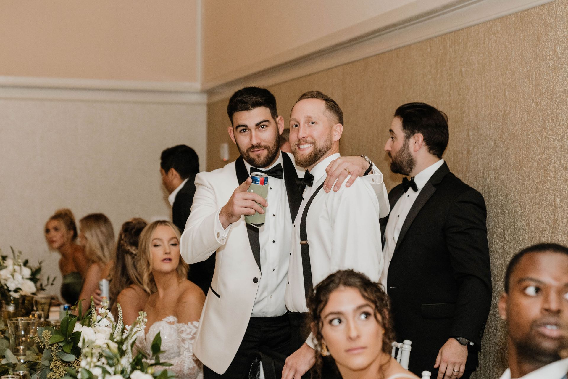 Men in tuxedos at wedding reception, one holding a drink.