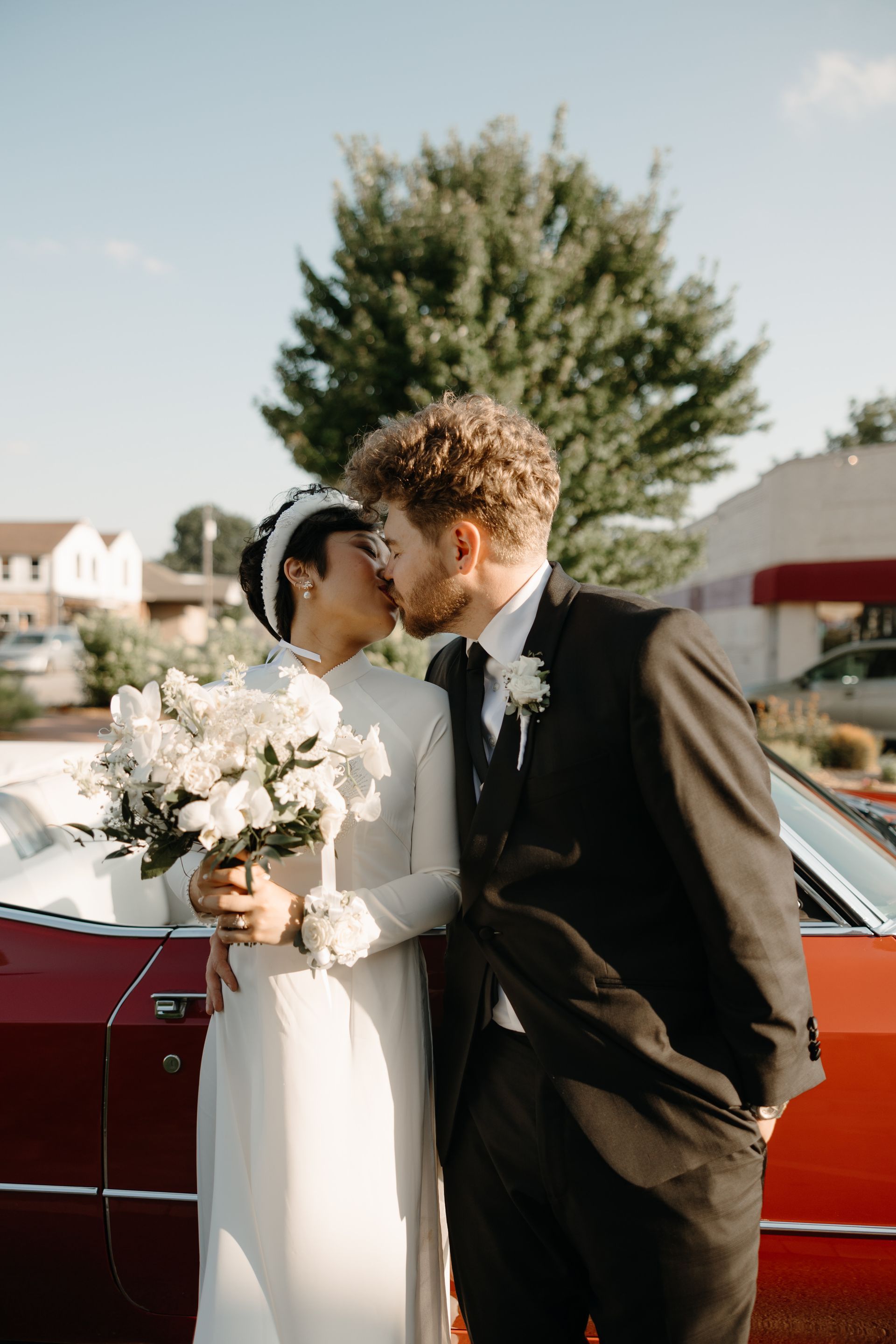 Couple kissing next to a red car, holding flowers. She wears a white dress, he wears a black suit, outside on a sunny day.