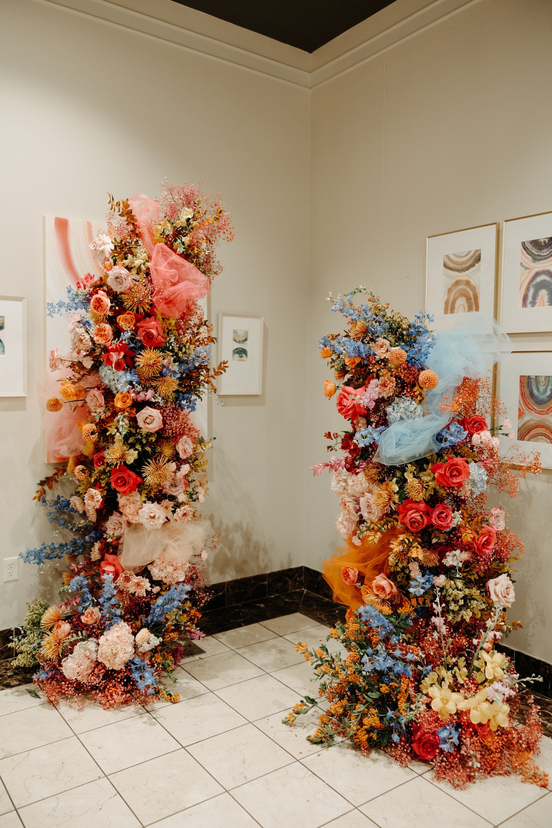 Two vibrant floral pillars with blue, pink, and orange flowers, tulle, in a gallery setting with art on walls.