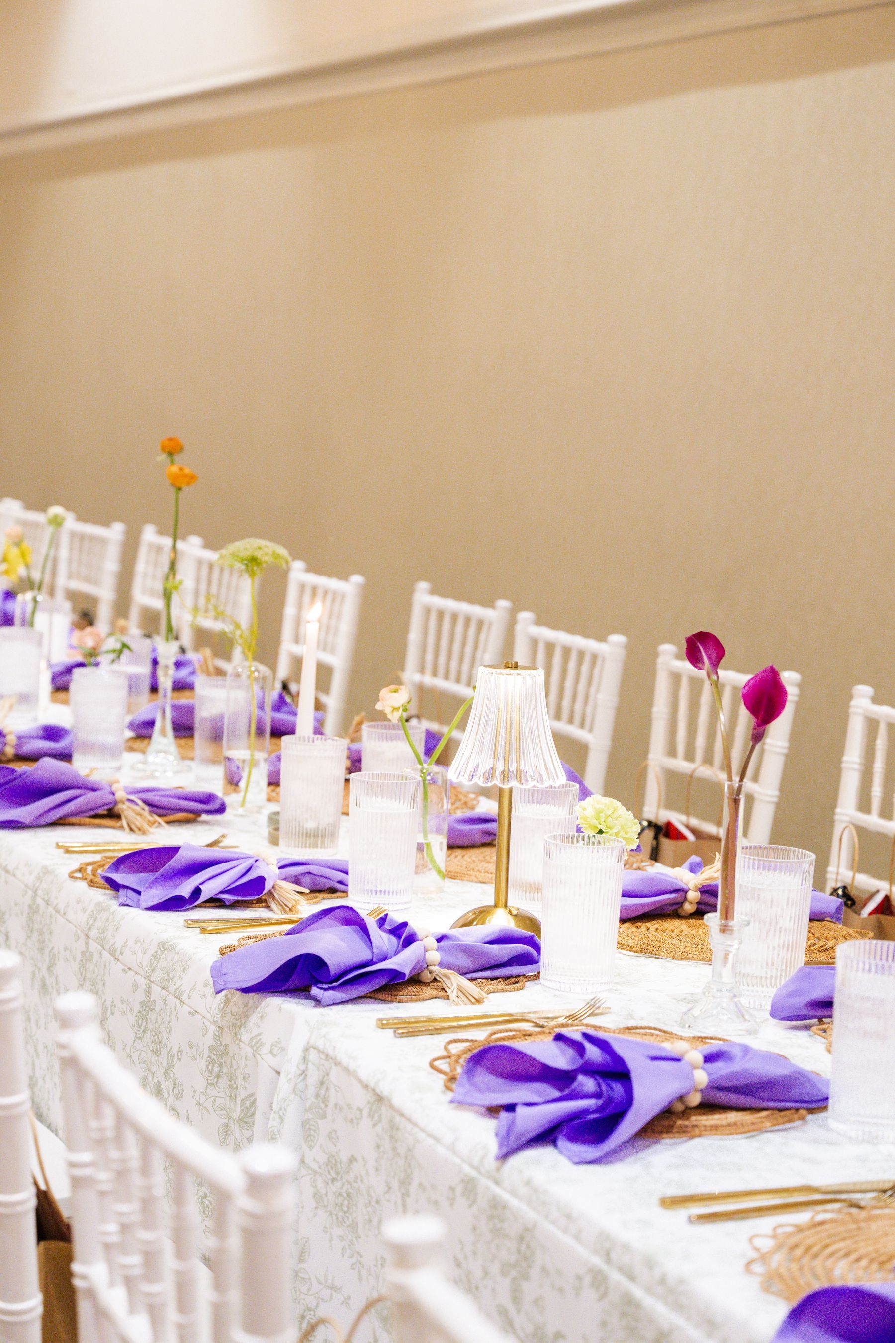 Long table set for a formal event, featuring purple napkins, gold accents, and white chairs.