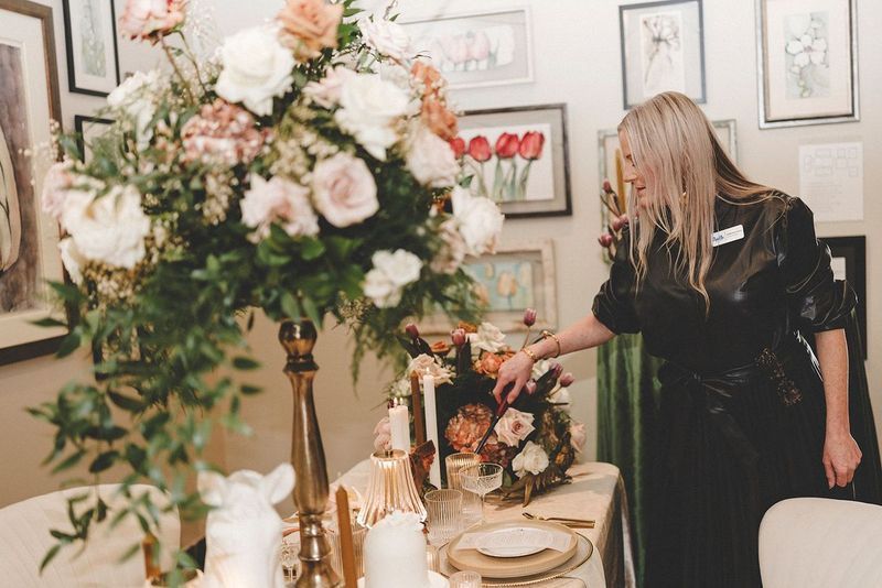Woman adjusting floral arrangement on a decorated table; art on the wall in the background.