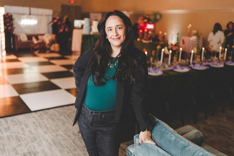 Woman in teal top and black blazer at an event, leaning on a teal sofa near a decorated table.