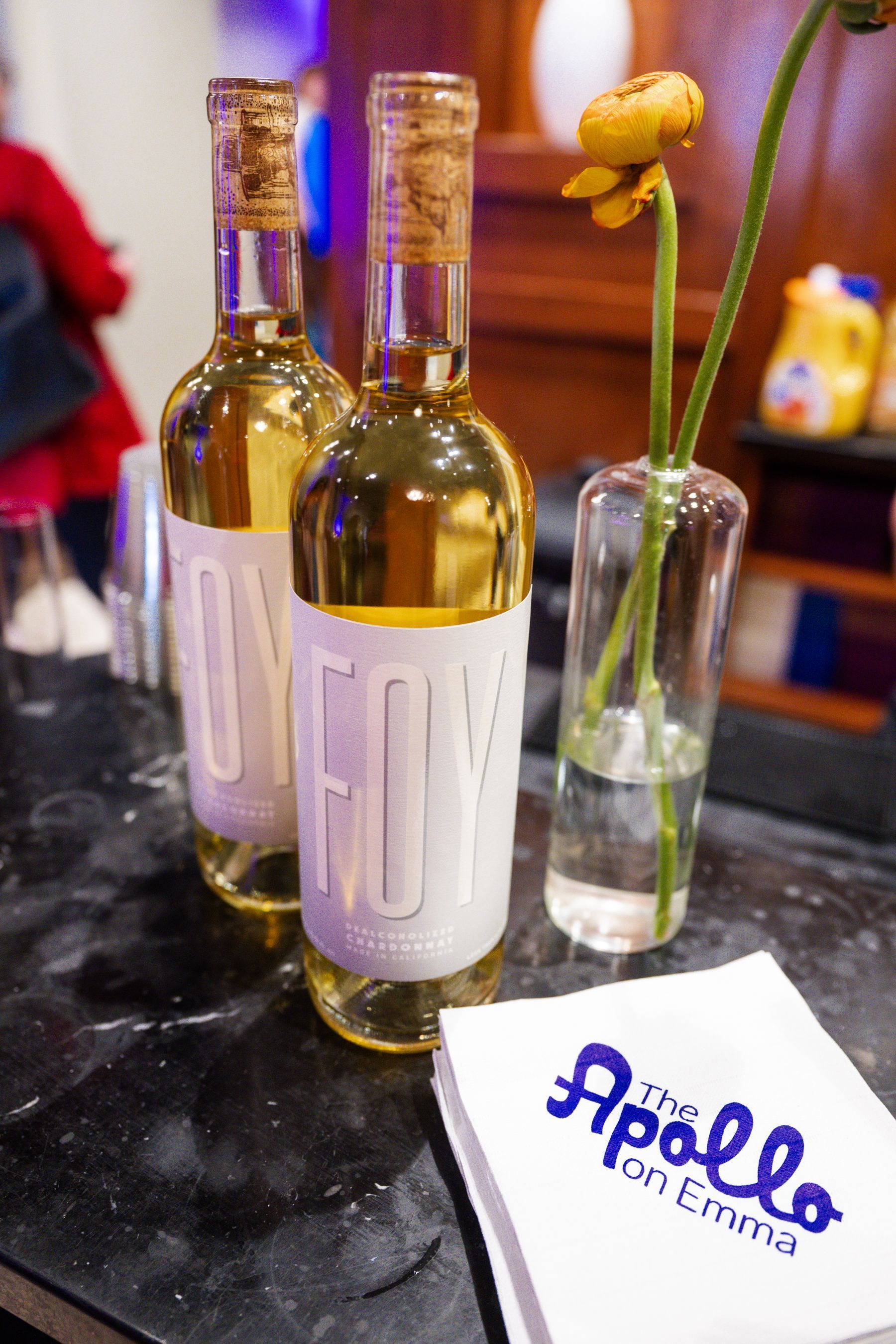 Two wine bottles with gold tops and white labels, flower in a vase, and a notebook with a logo.