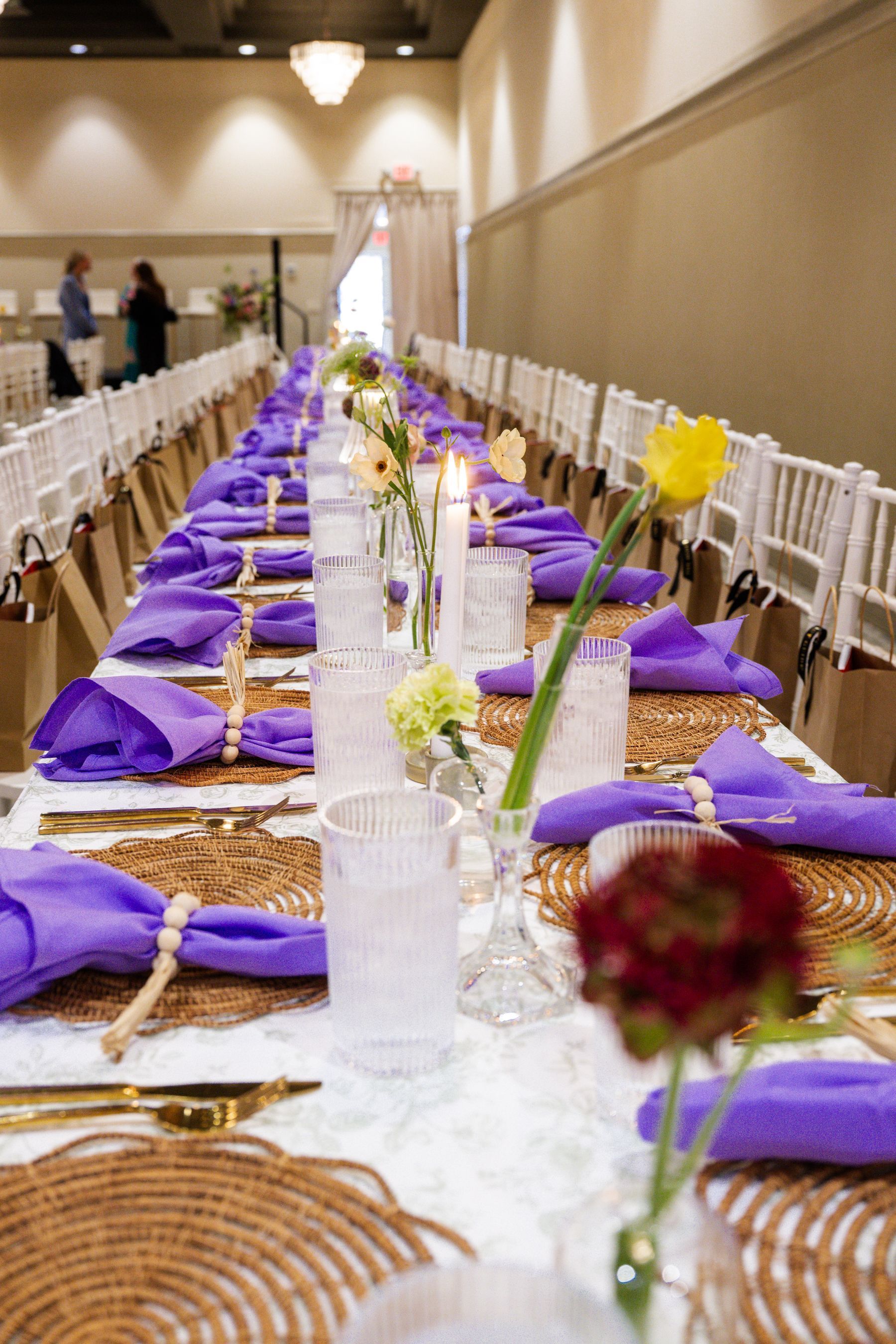 Long dining table set for event with purple napkins, floral centerpieces, and white chairs.