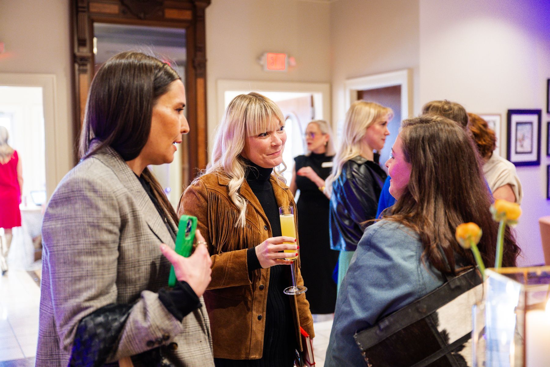 Group of people socializing indoors, some holding drinks. Well-lit room with art and a large mirror.