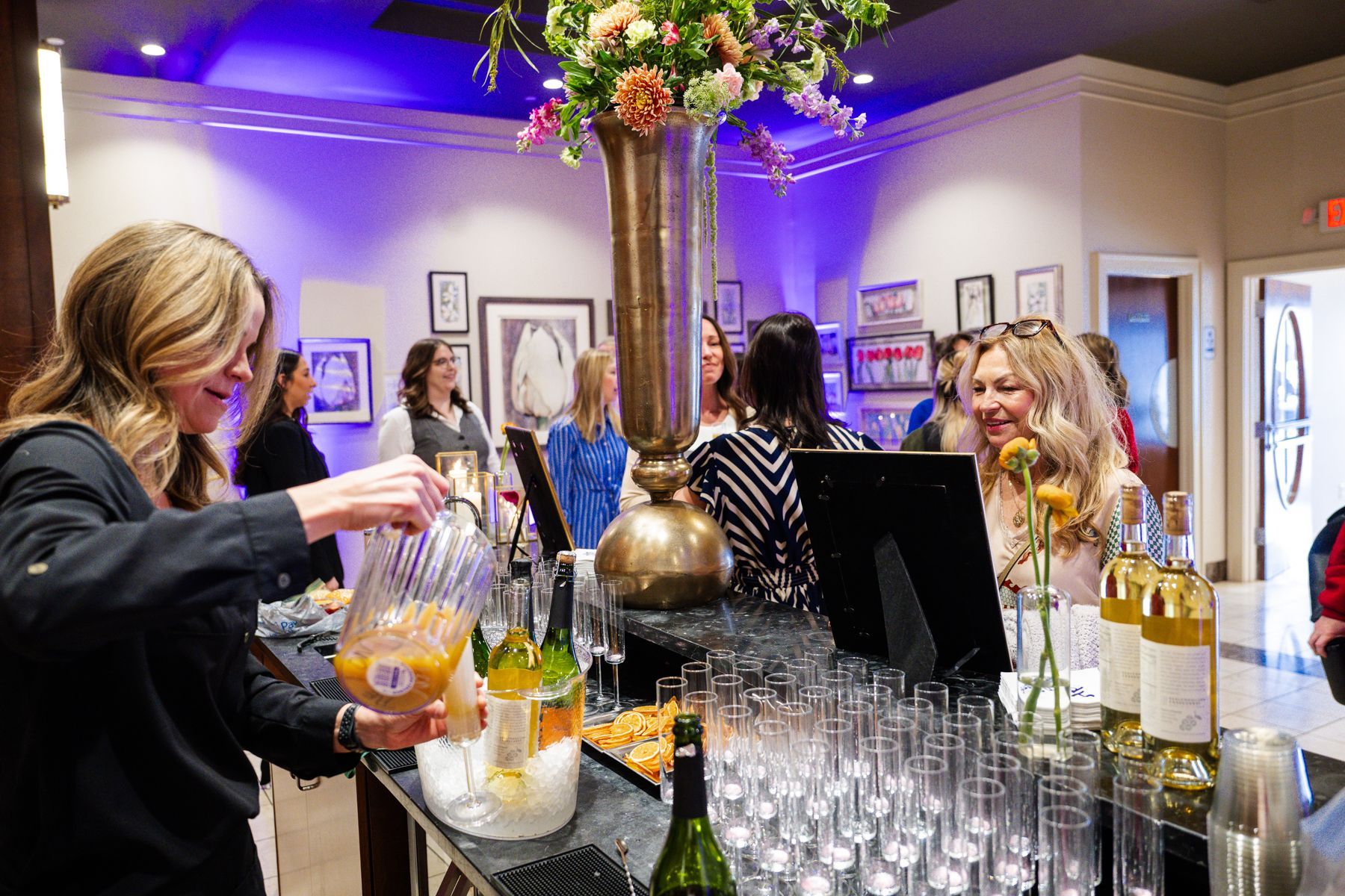 Woman pouring beverage at a bar in a well-lit event space; guests mingle in the background.