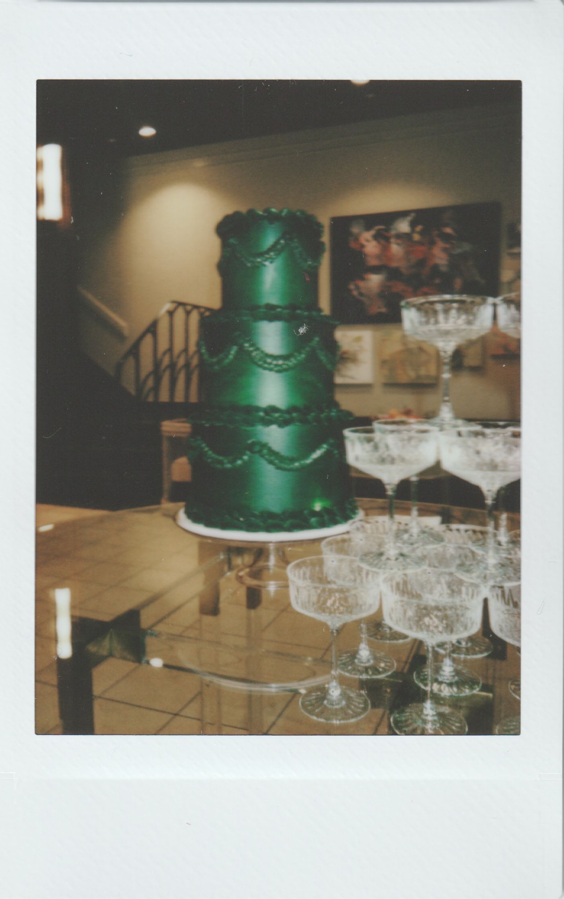 Three-tiered emerald green cake with garland detail, champagne glasses stacked on a glass table.