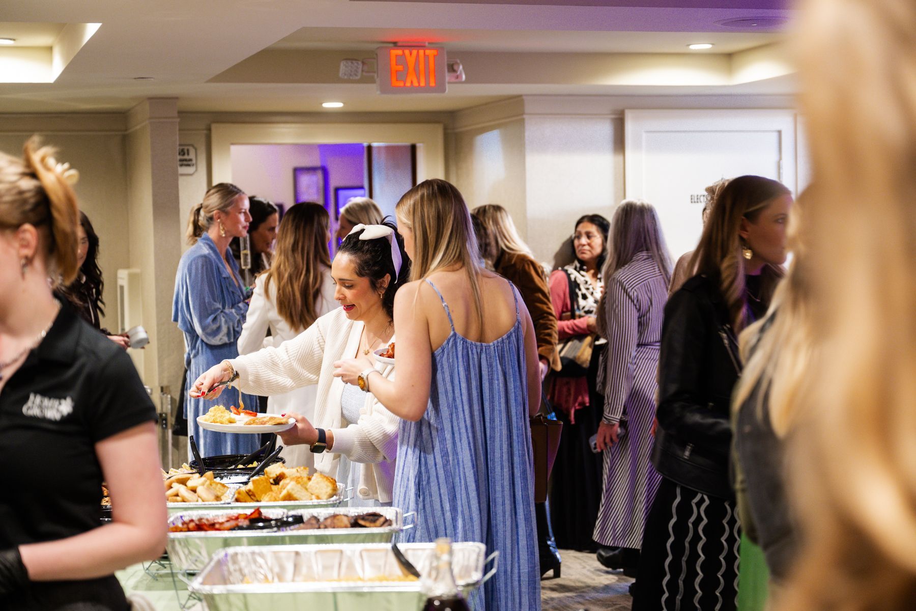 People at a gathering near a buffet, with food and drinks. An Exit sign is above a doorway.