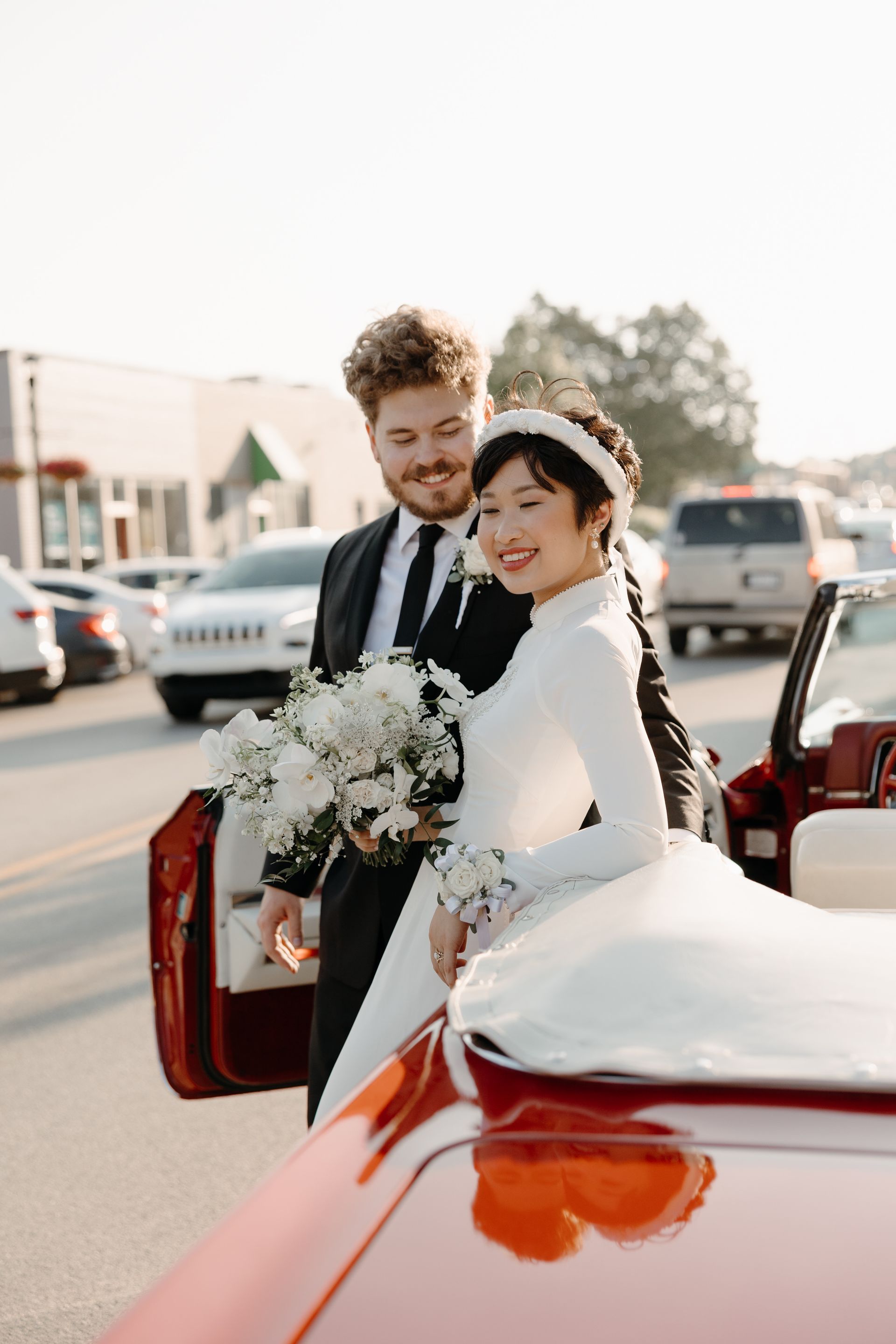 Wedding couple exiting a red car. Bride in white dress and headpiece, groom in a black suit. City street background.