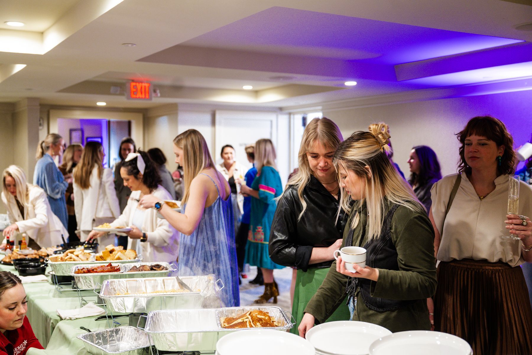 People at a buffet in a room, some serving themselves food.  A few hold plates and drinks.  Soft lighting.