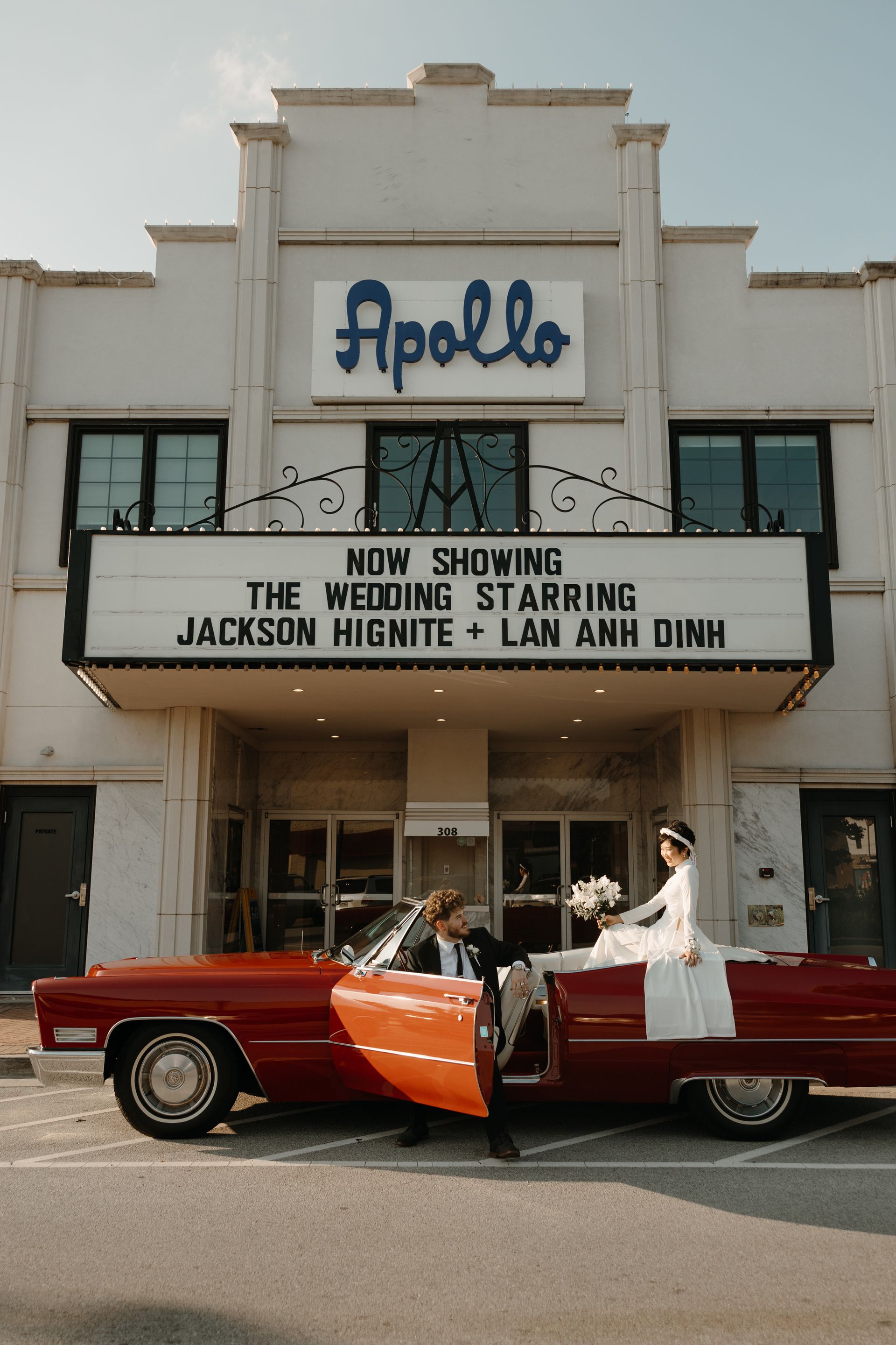 Newlyweds pose in front of a theater with their wedding date on the marquee, beside a light blue vintage car.