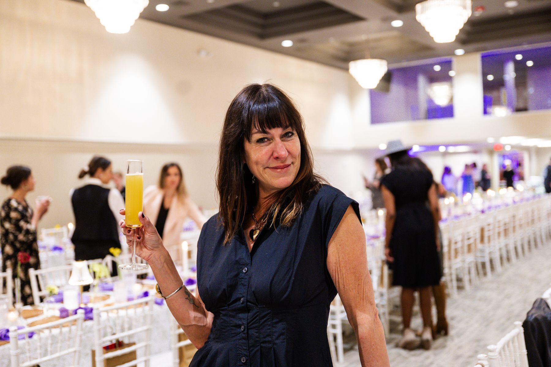 Woman smiling, holding a mimosa at a well-lit event. Tables with place settings in a large room.