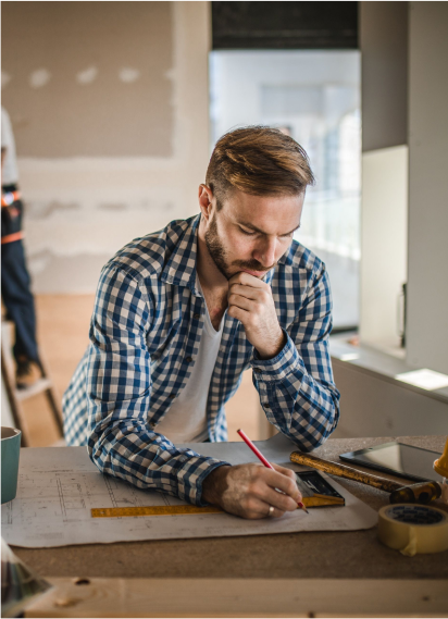 Man in plaid shirt sketches on blueprints at a construction site, hand to his chin, deep in thought.