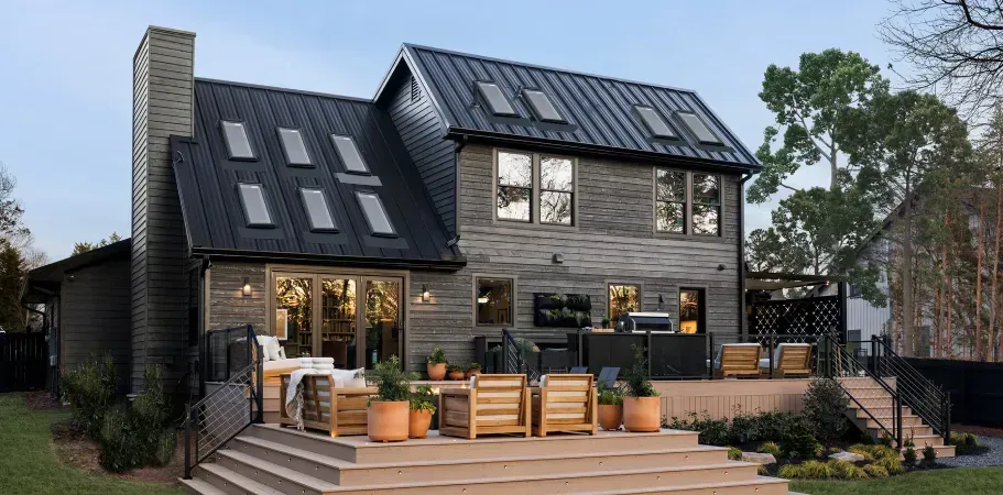 Back of a house with gray siding, black roof with skylights, and wooden deck with outdoor furniture.