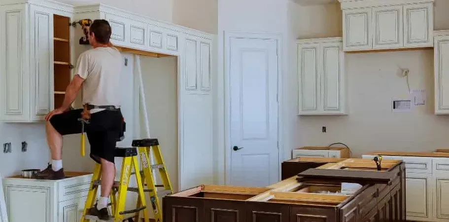A person is installing kitchen cabinets, standing on a step ladder, using a drill. The kitchen is under construction.