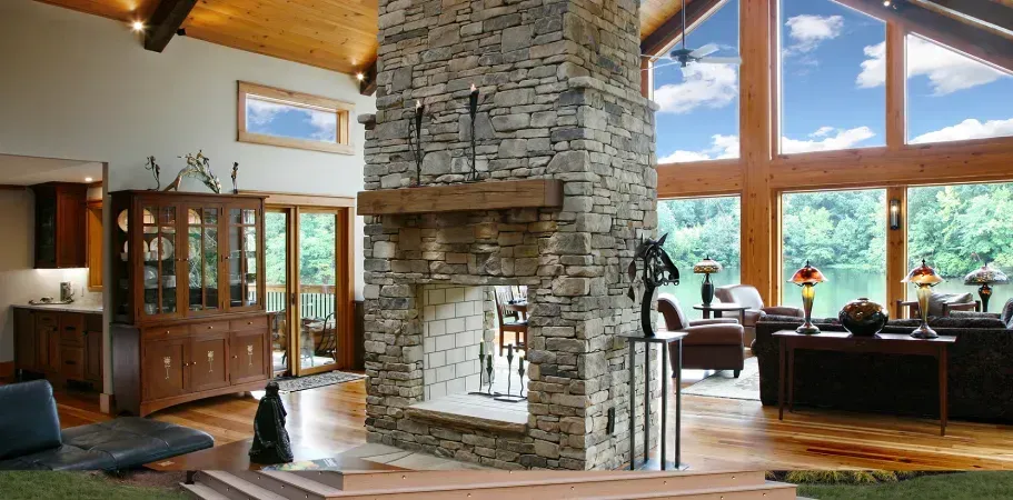 Living room with stone fireplace, wooden floor, and large windows with a view of a lake and blue sky.