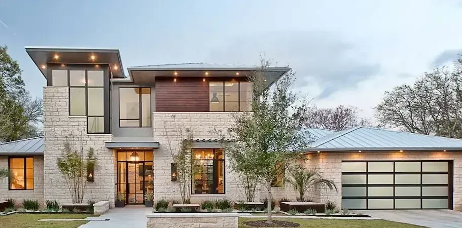 Modern, stone-clad house with large windows, a metal roof, and a glass-paneled garage door.