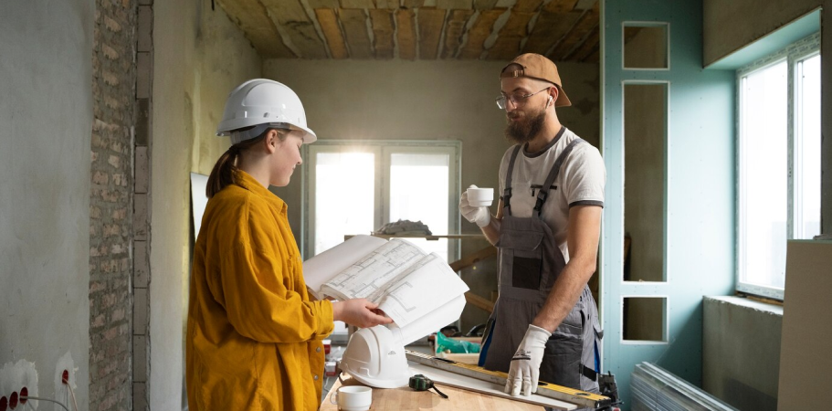 Two people in a construction site looking at blueprints, one with a coffee cup.