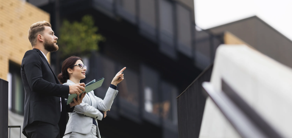Two people in suits looking at a building, one pointing, outdoors.