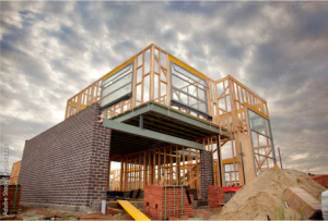 House under construction with wooden frame, brick wall, and steel beam. Cloudy sky in the background.