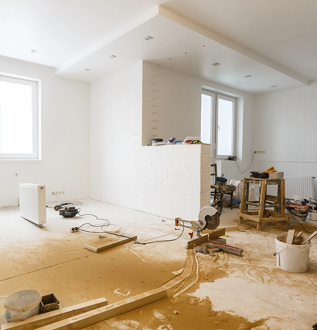Interior of a room under renovation; white walls, dusty floor with tools, saw, and a heater.