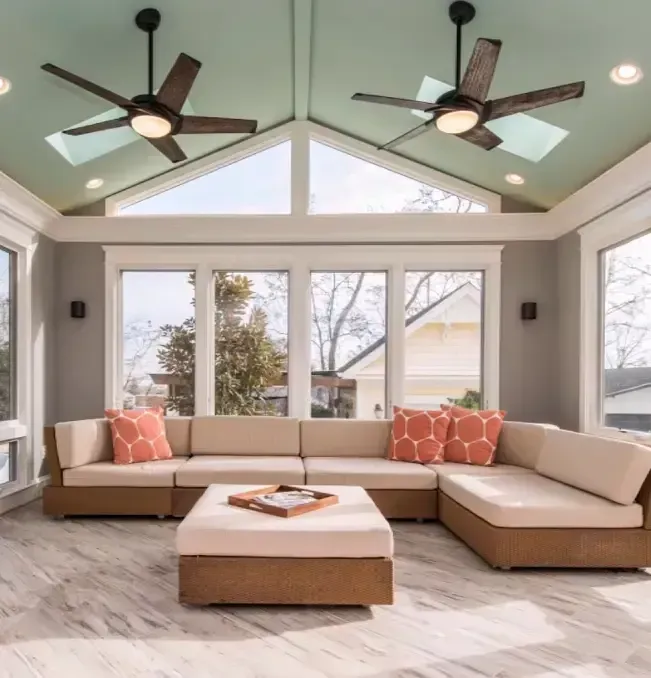 Sunroom with sectional sofa, orange pillows, ceiling fans, and large windows.