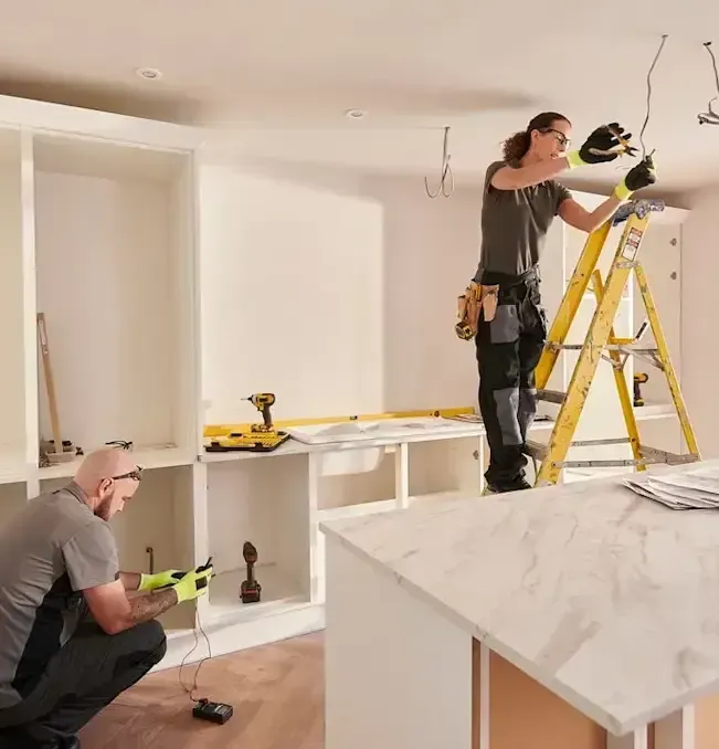 Two people renovating a kitchen: one on a ladder working on ceiling wiring, the other using a power tool on cabinets.