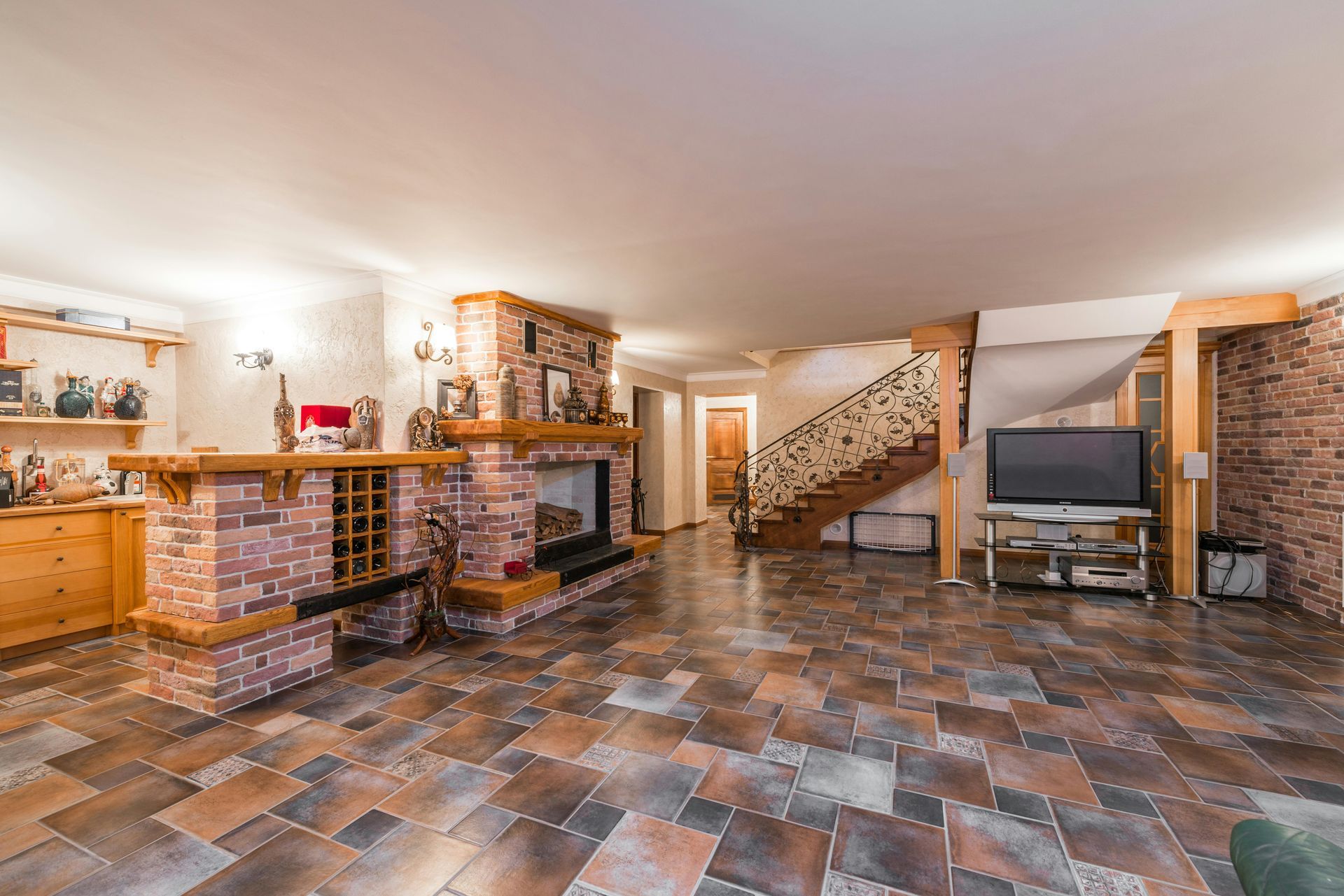 Spacious basement recreation room with tile floor, brick bar, TV, and staircase with decorative railing