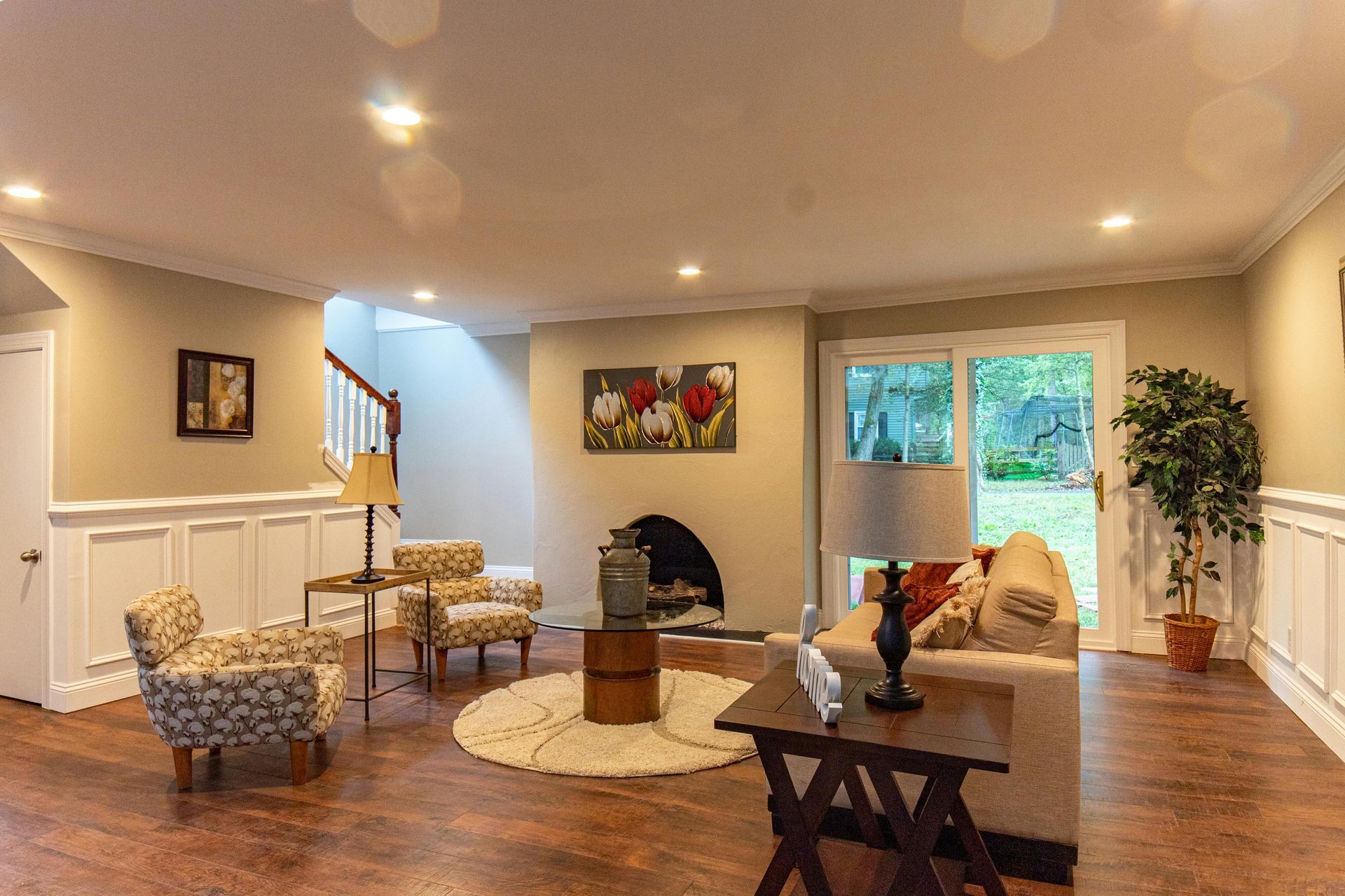 Bright living room with beige walls, sofa, chairs, coffee table, fireplace, and glass doors to a green yard