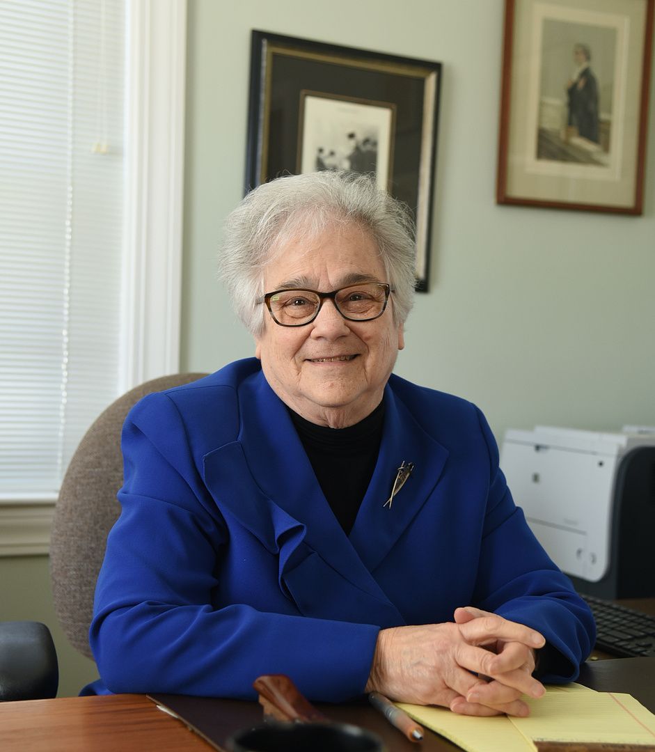 A woman in a blue jacket and glasses sits at a desk