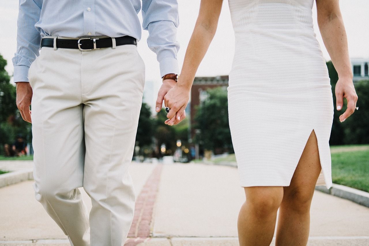 Couple holding hands walking on a path; the woman in a white dress, the man in a blue shirt and tan pants.