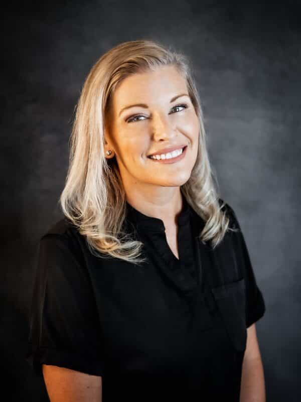 Blonde woman smiling, wearing black shirt, against dark background.