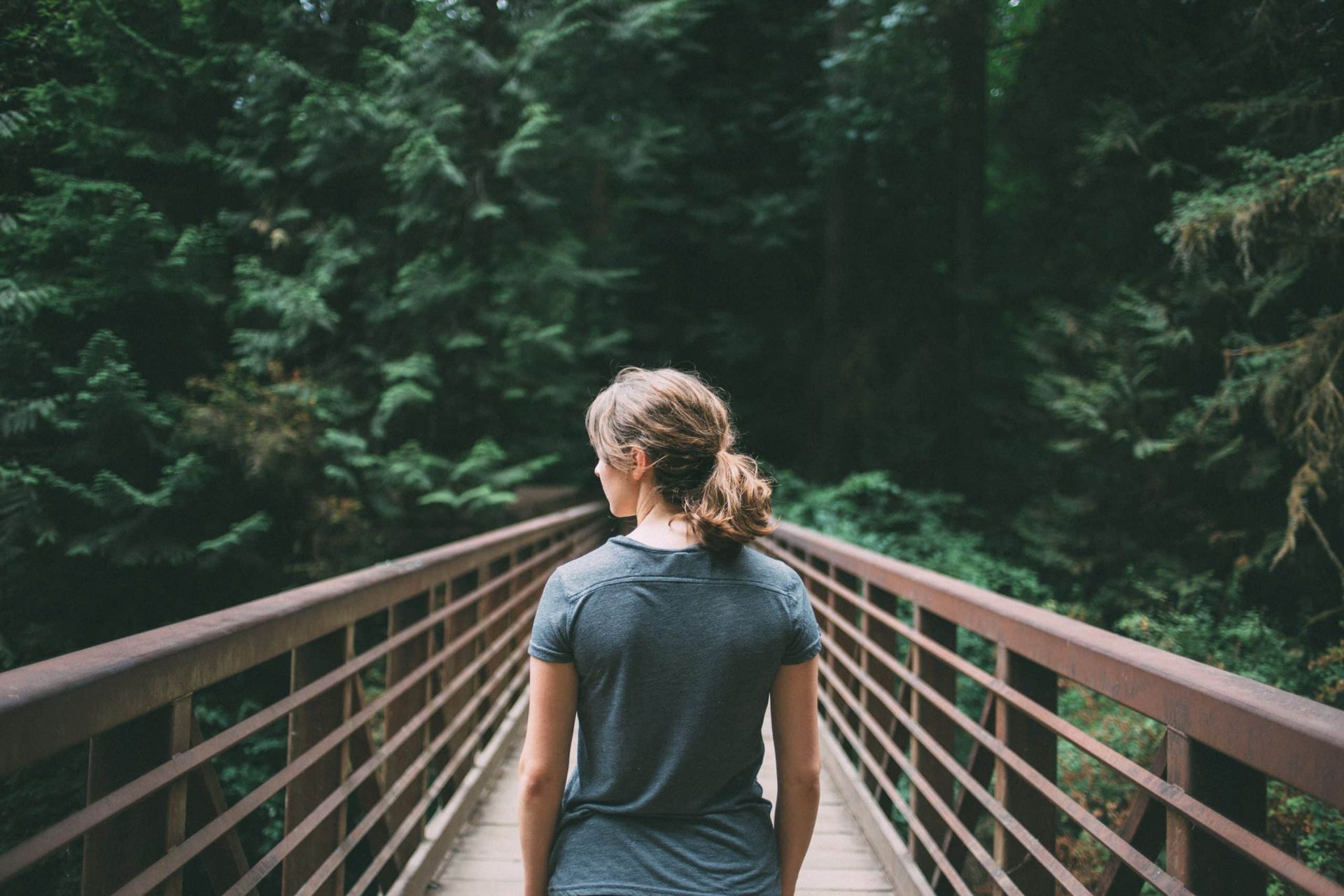 Woman walking on a wooden bridge in a forest; wearing a gray shirt, trees on either side.