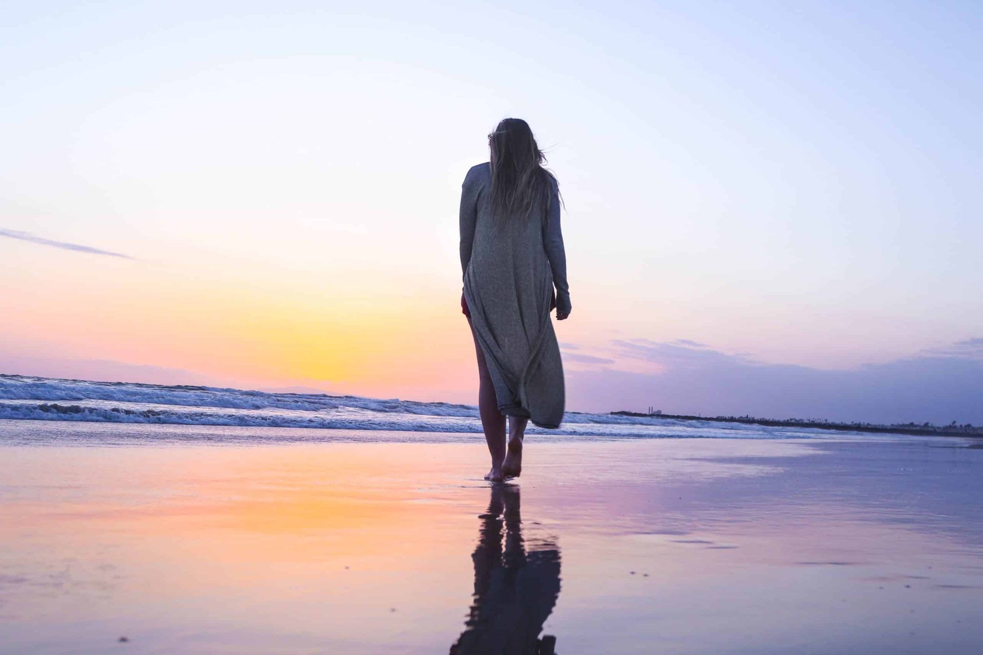 Woman walking on a beach at sunset, with reflection in wet sand, blue and orange sky.