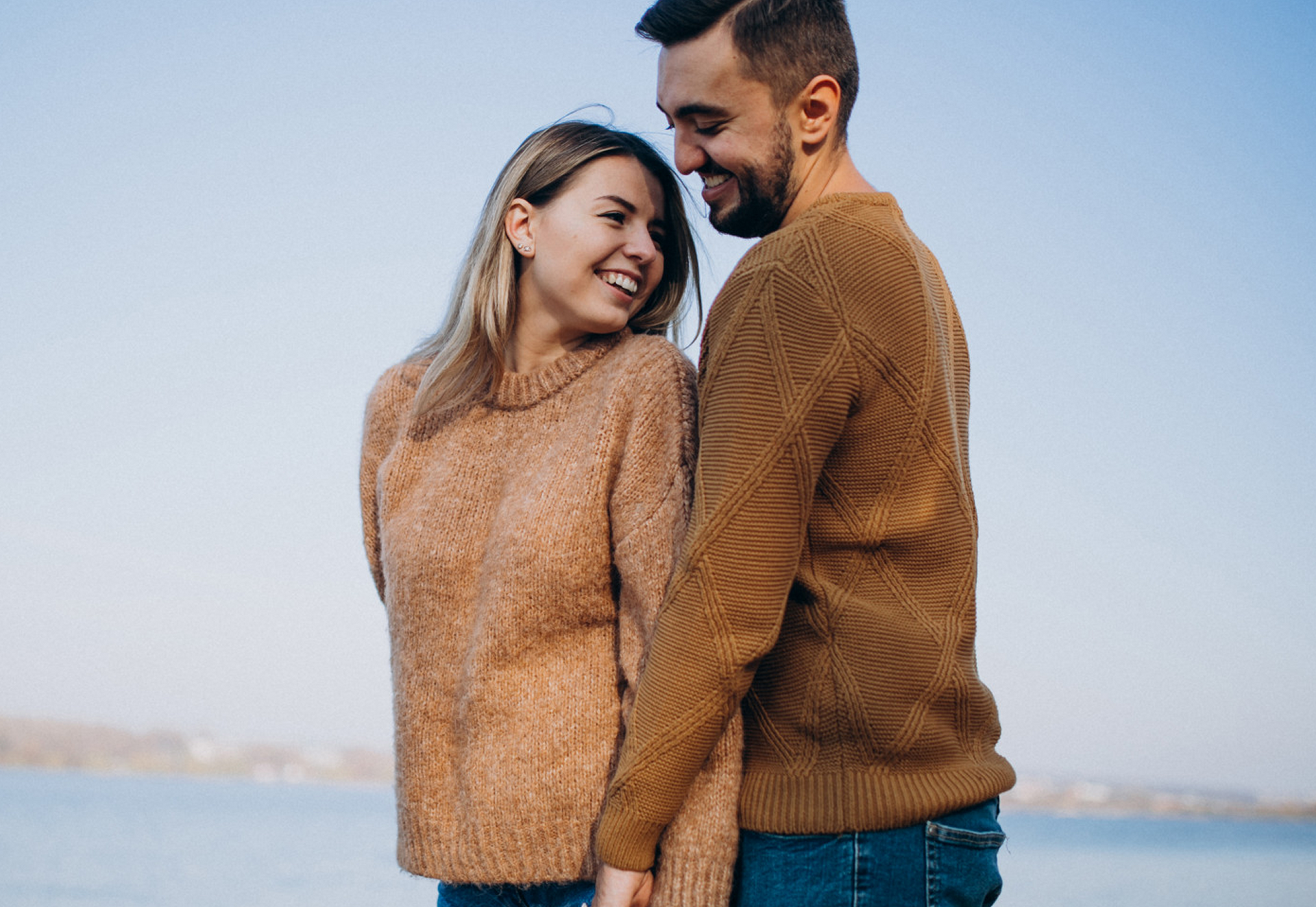 Smiling couple in matching brown sweaters holding hands by water.
