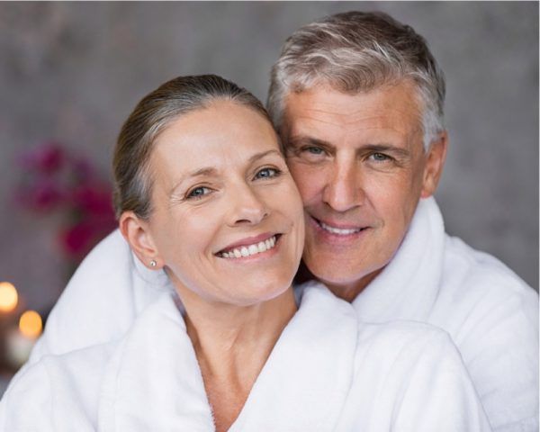 Smiling couple in white robes, embracing. Spa-like setting.