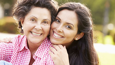 Mother and daughter smiling outdoors; mother with short curly hair, daughter with long dark hair.