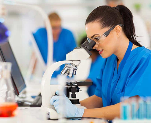 Woman in lab coat and glasses looks into microscope in a lab.