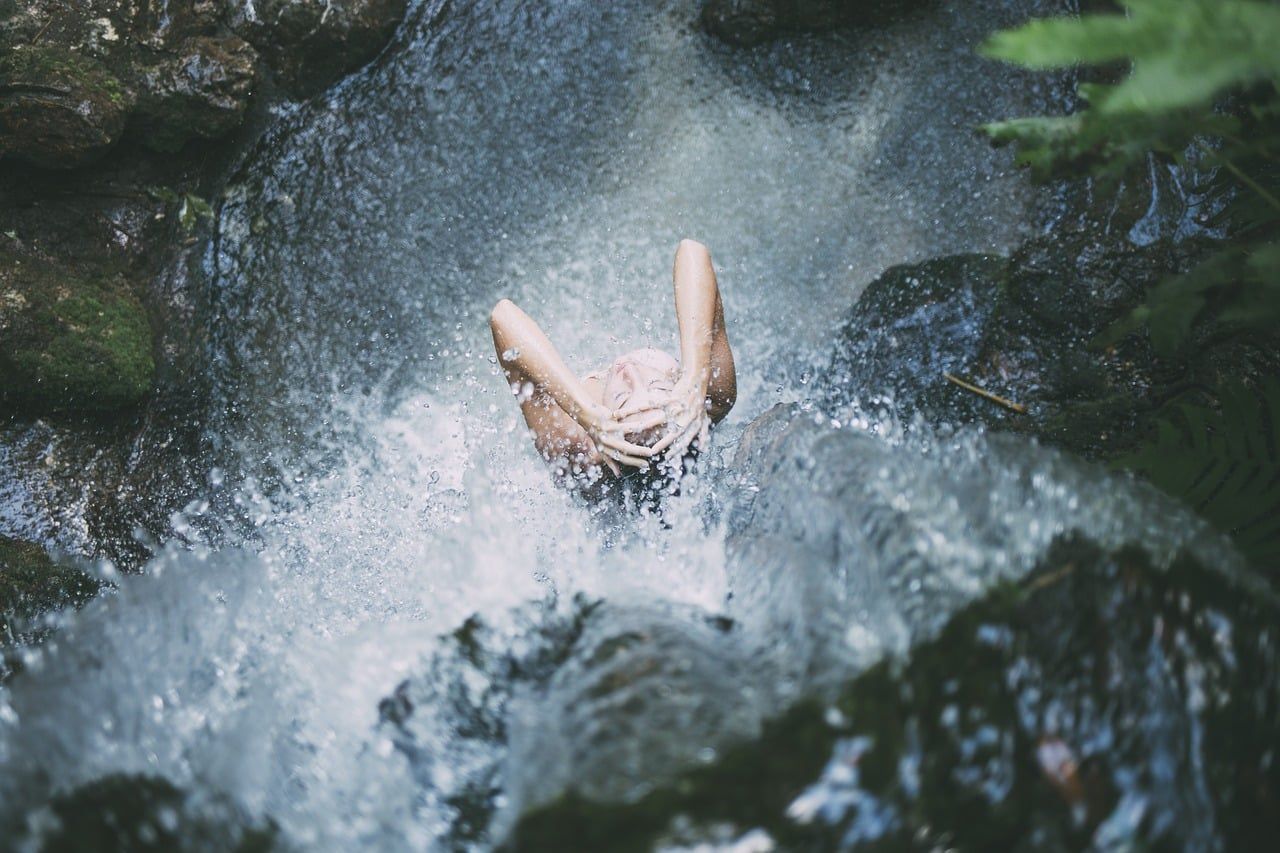 Person submerged in rushing water, arms raised, in a rocky, wooded area.