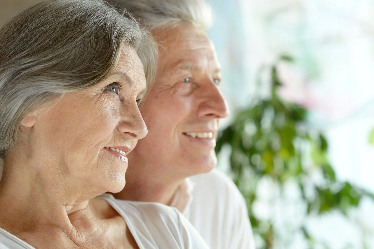 Older couple, side-by-side, looking outside, smiling. Light-colored clothing, gray hair, bright background.