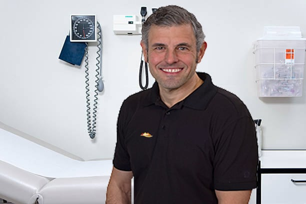 Man in black shirt smiles in a doctor's office, next to a blood pressure monitor and examination table.