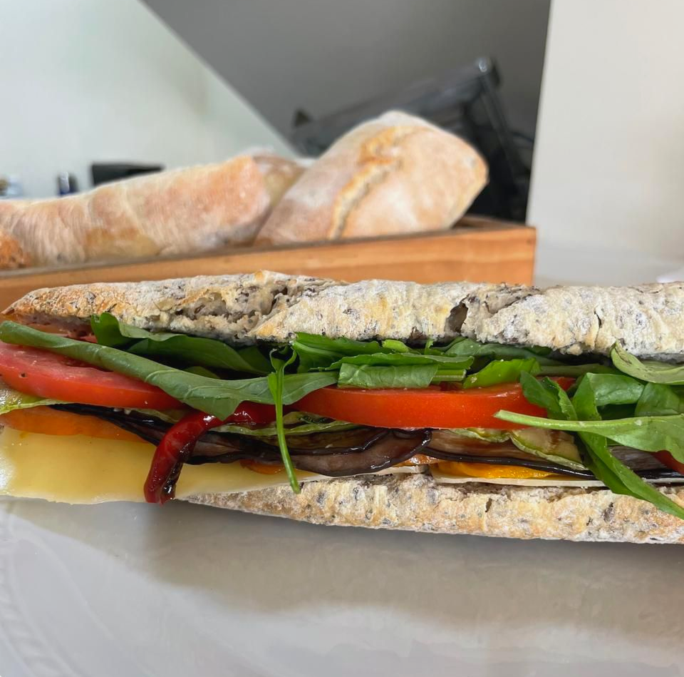 Un bocadillo de baguette con semillas, queso, tomate y verduras de hoja verde en un plato blanco, con pan extra al fondo.