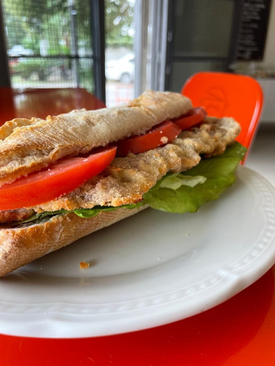 Un sándwich de milanesa de pollo empanizada con lechuga y rodajas de tomate en un plato blanco sobre un fondo naranja.