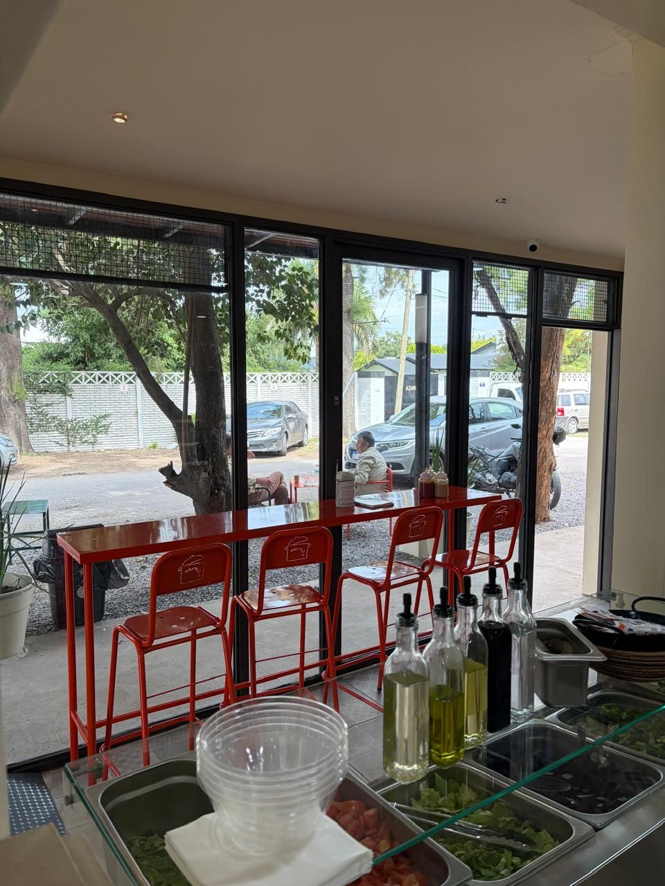 Taburetes de metal rojo en una barra frente a grandes ventanales, con botellas de condimentos y bandejas para preparar alimentos en primer plano.