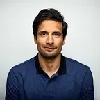 Man with dark hair and a blue polo shirt smiles in front of a white background.