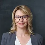 Woman with blonde hair, glasses, wearing a gray blazer smiles against a dark background.