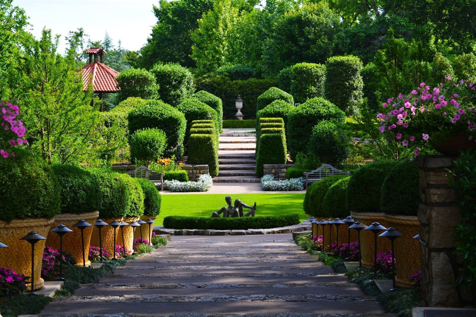 Stone path leads to a manicured garden with topiary shrubs and steps. Lush greenery, flowers, and a fountain.